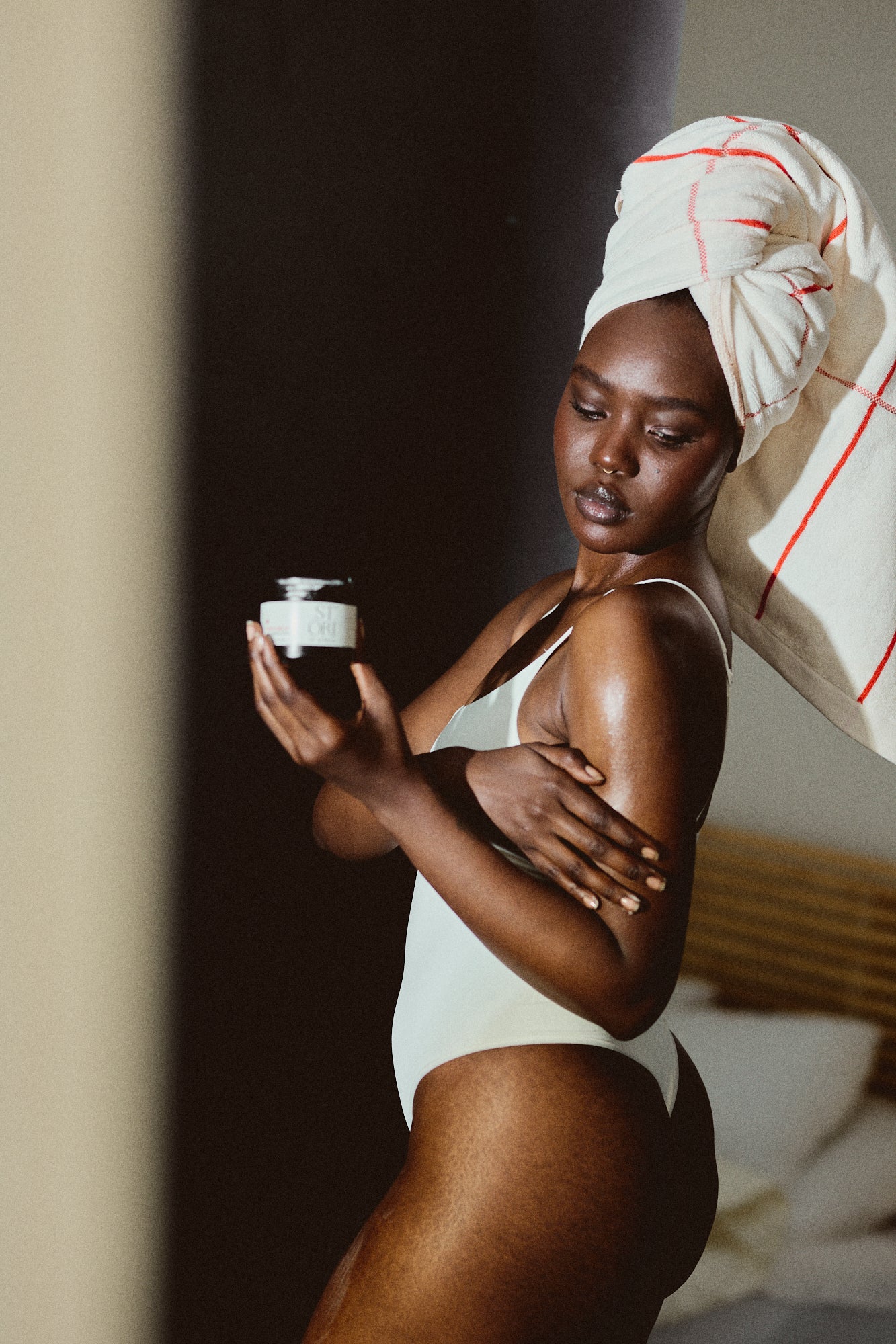 Black woman in a white swimsuit with a towel on her head, holding a jar of Stori of Africa Shea Balm and moisturising her skin. 